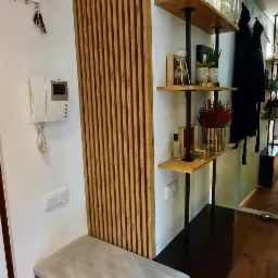 Nowoczesny przedpokój z drewnianą ścianką działową z listew, siedziskiem z szarego materiału i półkami na czarnych metalowych wspornikach, dekoracje na półkach, drewniana podłoga.