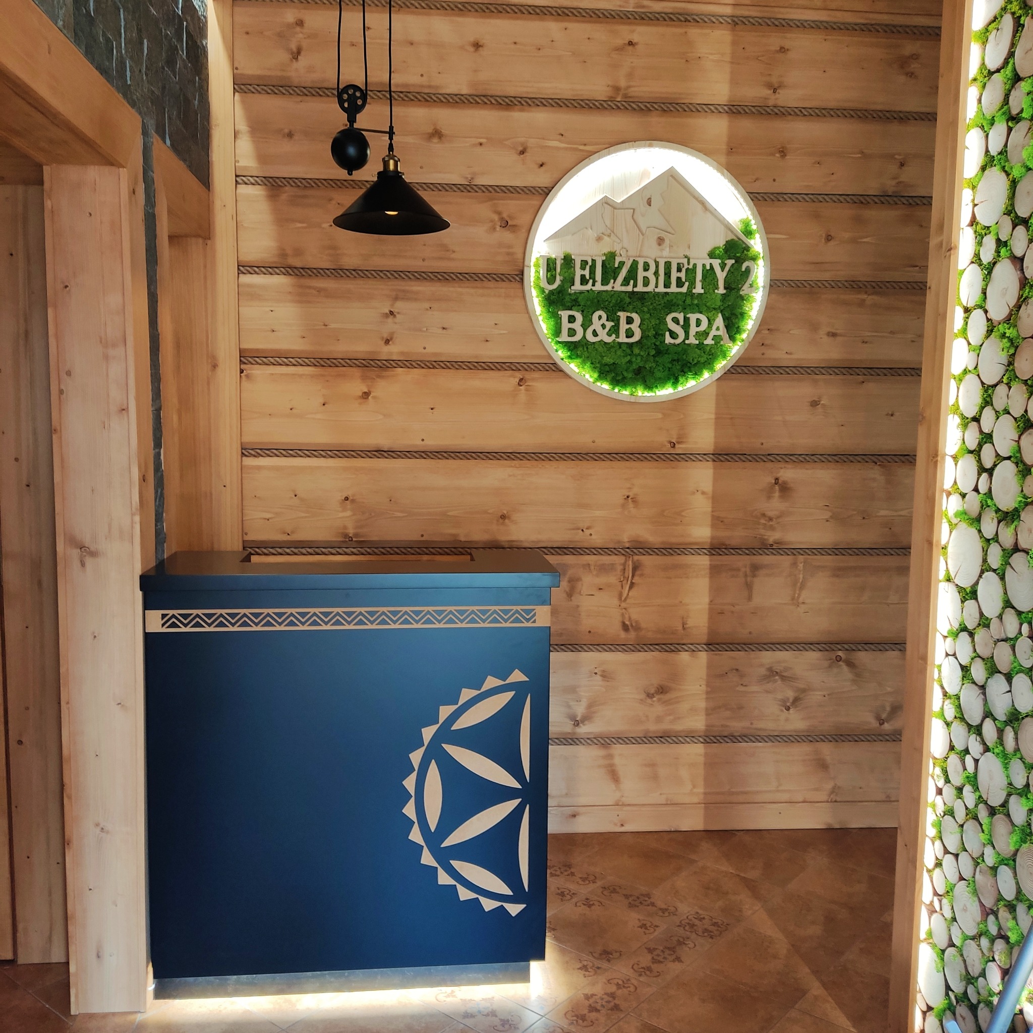 Recepcja w stylu góralskim w Blok dobryszyce: drewniana ściana z dekoracyjnym sznurem, logo B&B SPA z mchu, niebieska lada z ludowym wzorem i podświetleniem.