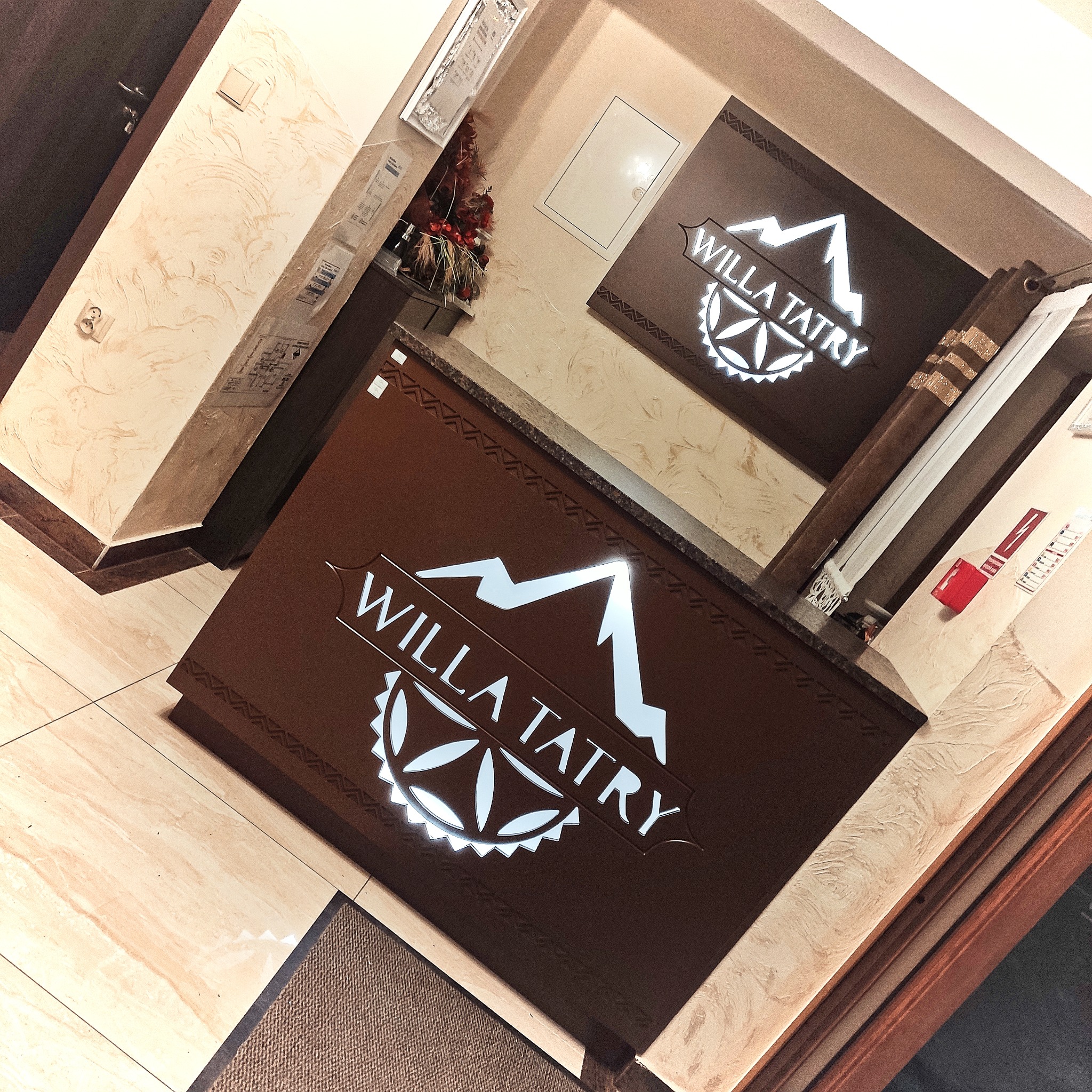 Recepcja hotelowa z brązowym frontem i podświetlanym logo 'Willa Tatry' w kształcie gór i stylizowanego kwiatu.