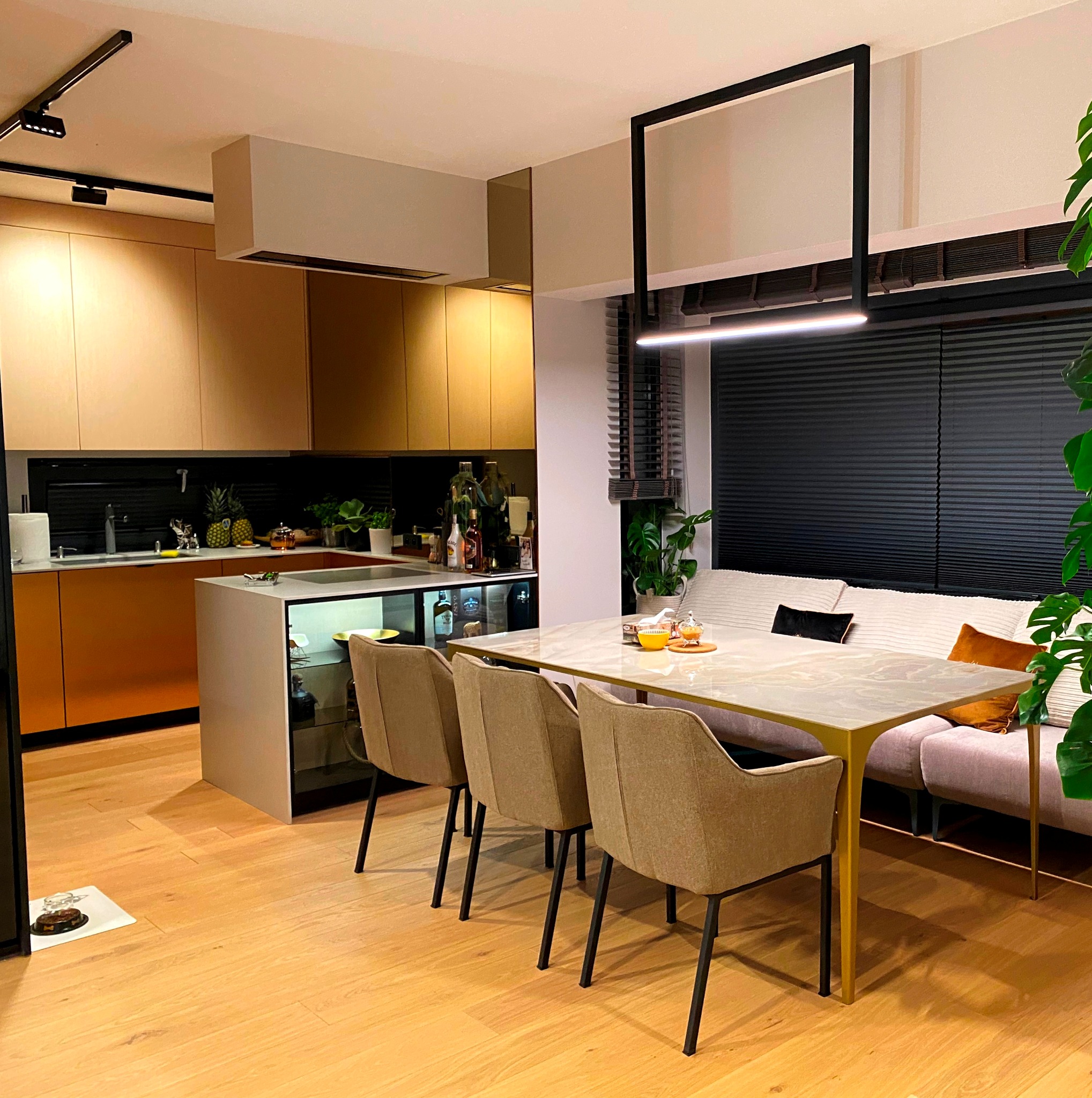 Nowoczesna jadalnia z blatem imitującym marmur, otoczona krzesłami z beżową tapicerką, z widokiem na kuchnię z frontami w odcieniach brązu i złotego, oświetlona lampą w kształcie czarnej ramy.
