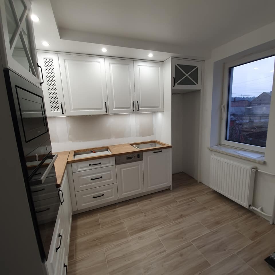 Wnętrze kuchni w trakcie montażu, z białymi szafkami z czarnymi uchwytami, drewnianym blatem bez zamontowanej płyty grzewczej i zlewu, oraz widocznymi śladami prac wykończeniowych na ścianie.