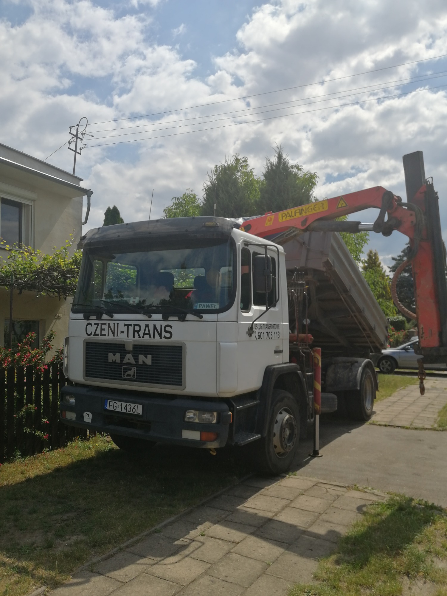Biała ciężarówka MAN z wywrotką i dźwigiem Palfinger, zaparkowana na chodniku obok domu z zielonymi pnączami, widoczne logo firmy transportowej CZENI-TRANS.
