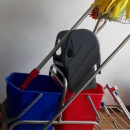 Wózek do sprzątania z dwoma wiadrami: niebieskim i czerwonym, oraz wyciskarką i mopem z żółtą końcówką, stojący na drewnianej podłodze.