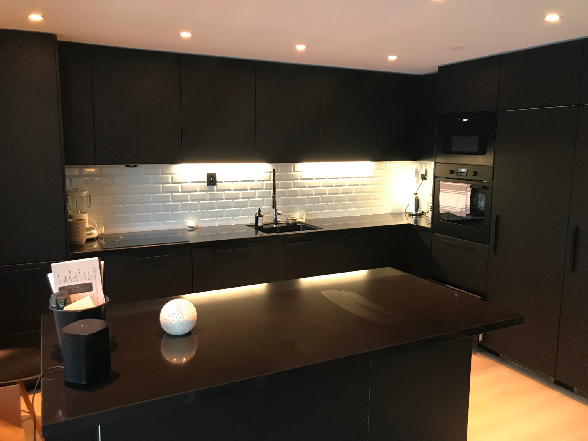 Nowoczesna, czarna kuchnia z wyspą, białymi płytkami metra i oświetleniem LED pod szafkami.