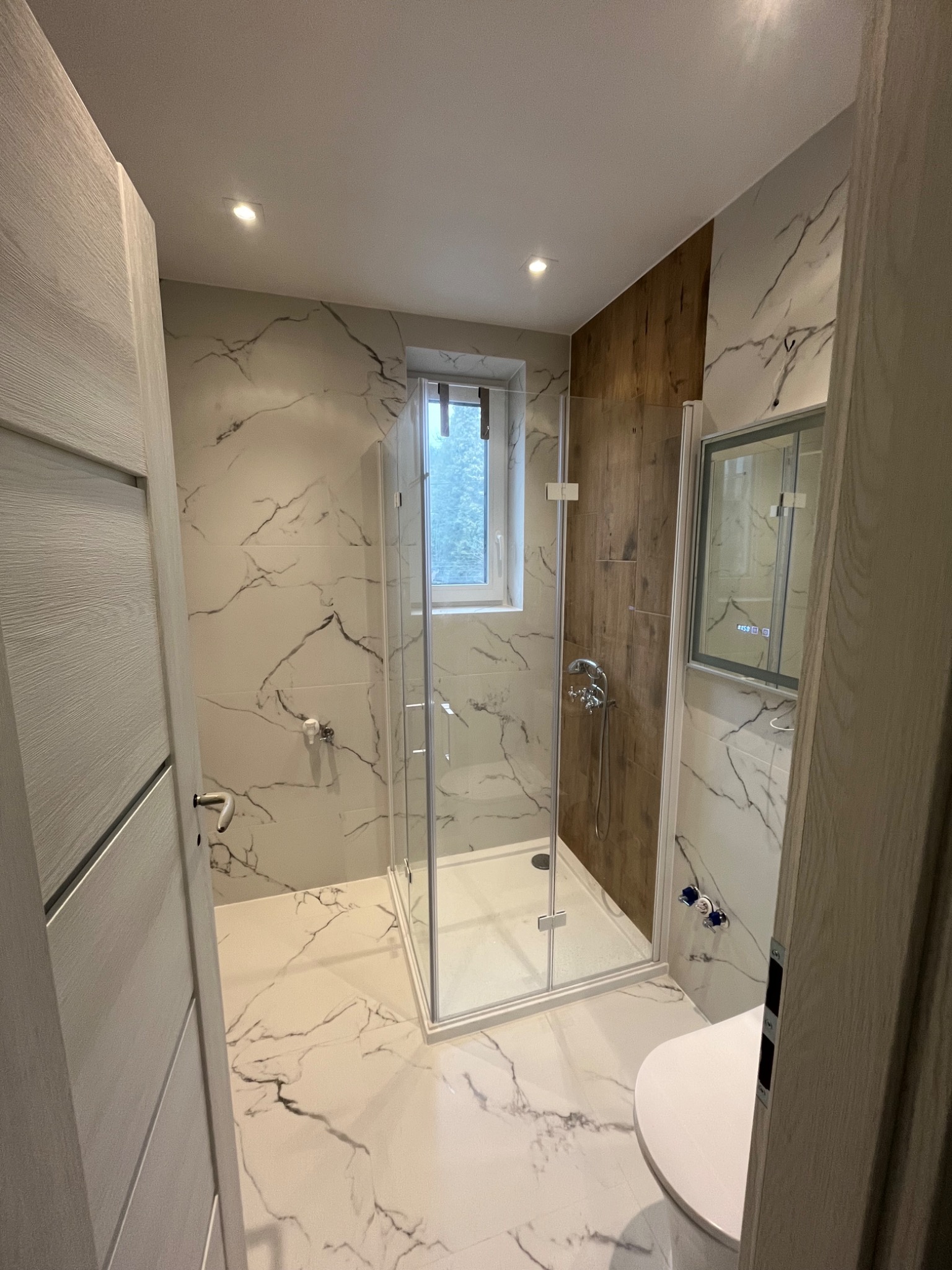Nowoczesna łazienka po remoncie: kabina prysznicowa ze szklanymi drzwiami, marmurowe płytki na ścianach i podłodze, drewniana ściana, widok zza uchylonych drzwi.