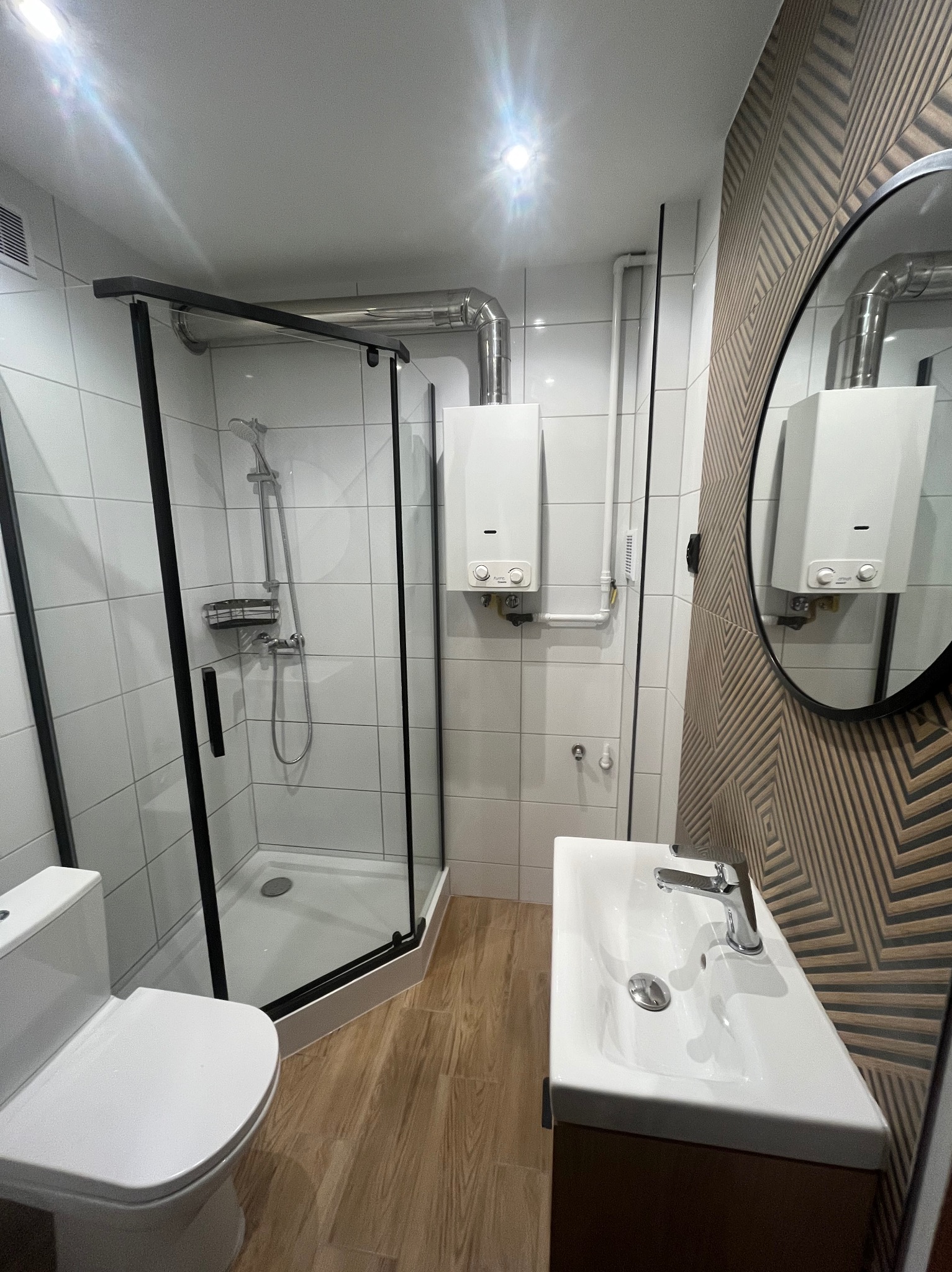 Nowoczesna łazienka po remoncie w Sosnowcu: kabina prysznicowa z czarnymi ramami, umywalka z szafką, dekoracyjna ściana i lustro. Widoczny bojler.