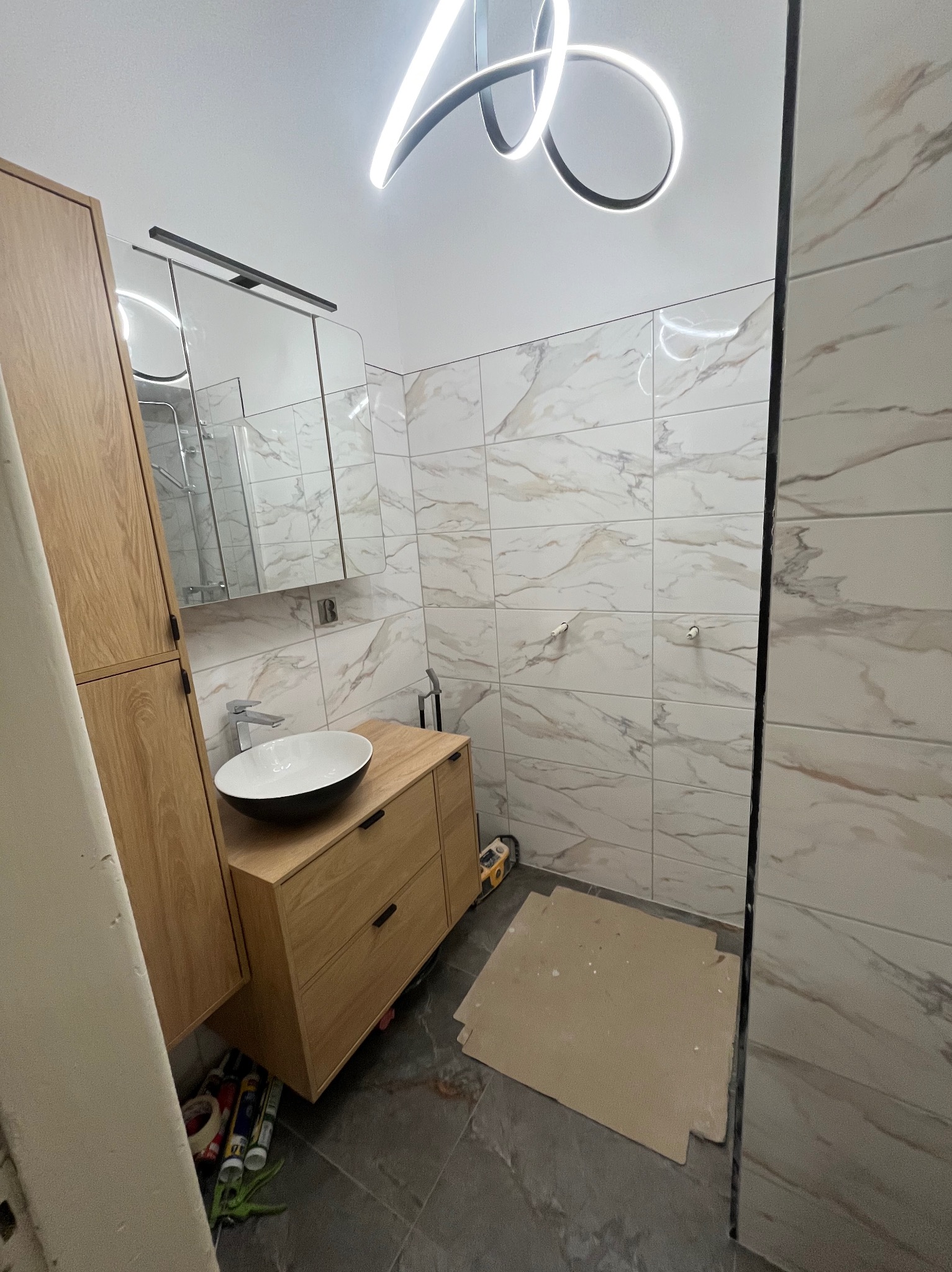 Nowoczesna łazienka w trakcie wykończenia: drewniana szafka z umywalką, lustro z oświetleniem, marmurowe płytki na ścianach, designerska lampa sufitowa.