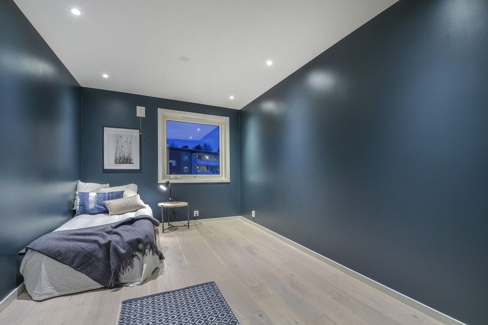 Pokój z gładkimi, granatowymi ścianami, jasną drewnianą podłogą i minimalistycznym wystrojem, w tym małym łóżkiem z dekoracyjnymi poduszkami i pledem, oraz dywanem w geometryczny wzór.