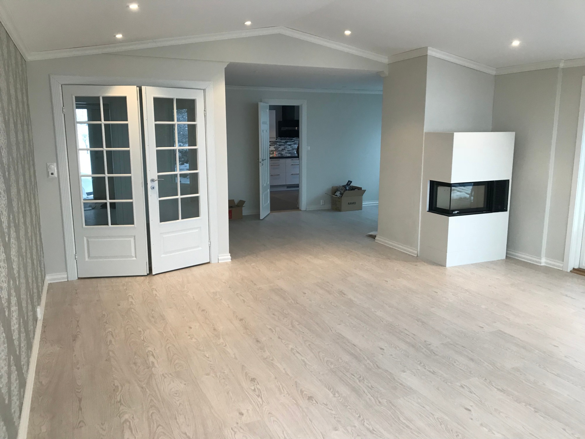 Jasne panele podłogowe w przestronnym salonie z białymi, przeszklonymi drzwiami dwuskrzydłowymi, minimalistycznym kominkiem i widokiem na kuchnię w tle.