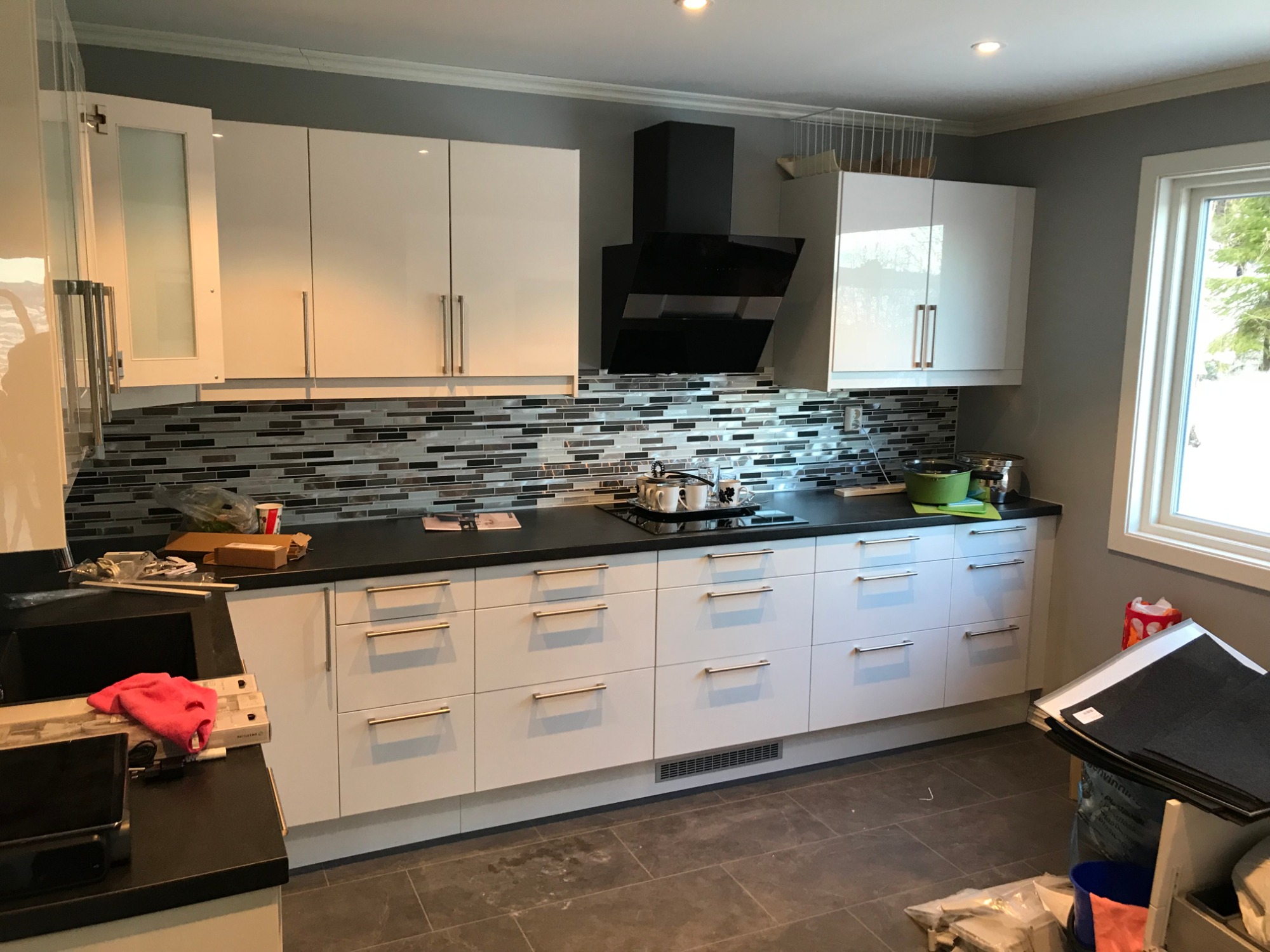 Nowoczesna kuchnia z białymi szafkami, czarnym blatem i mozaiką na ścianie, widoczne garnki na płycie grzewczej i akcesoria kuchenne, podłoga wyłożona szarą płytką.