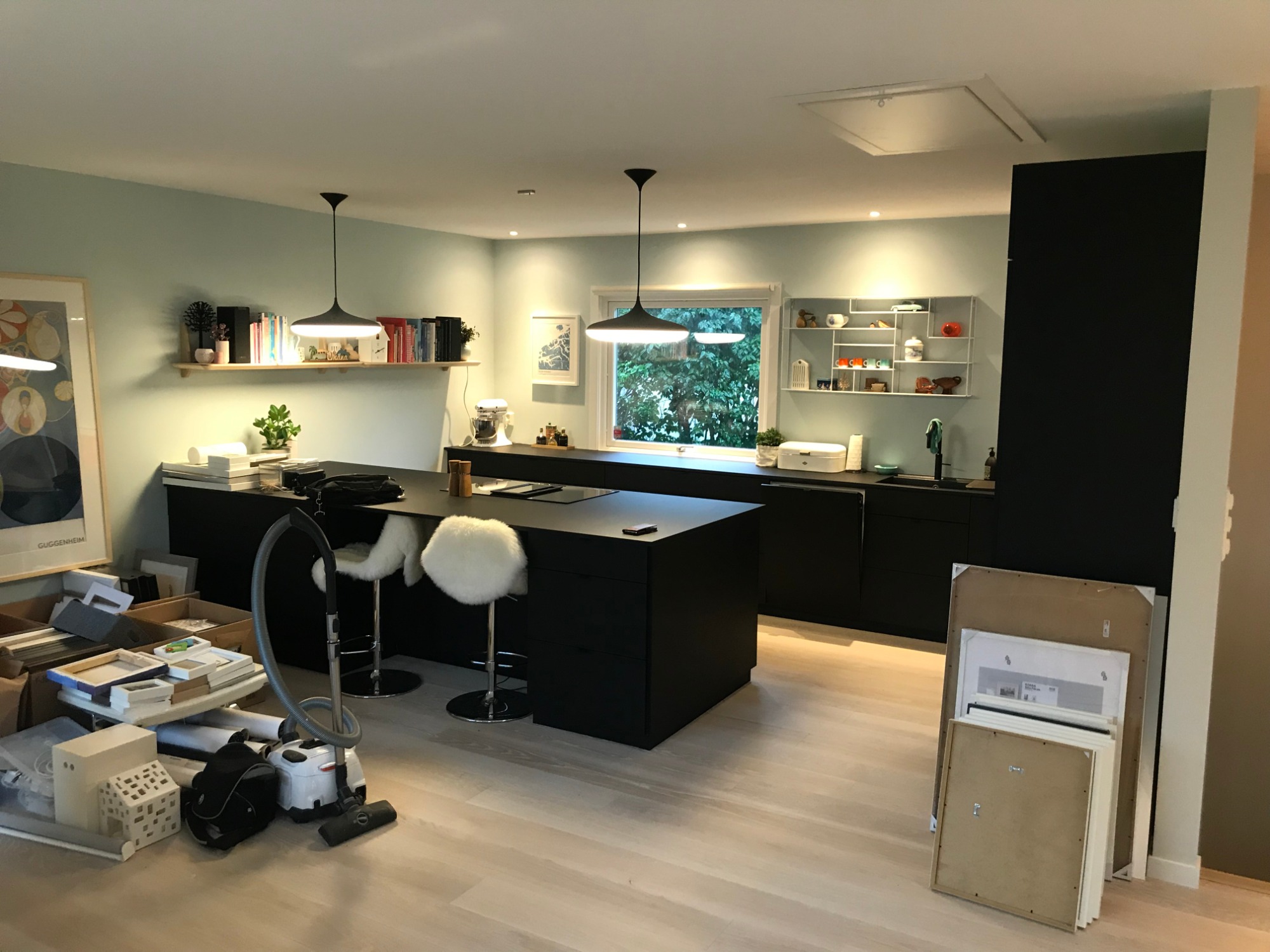 Jasne panele podłogowe w nowocześnie urządzonej kuchni z czarną wyspą i białymi hokerami; widoczny odkurzacz i kartony z ramkami, sugerujące prace wykończeniowe.