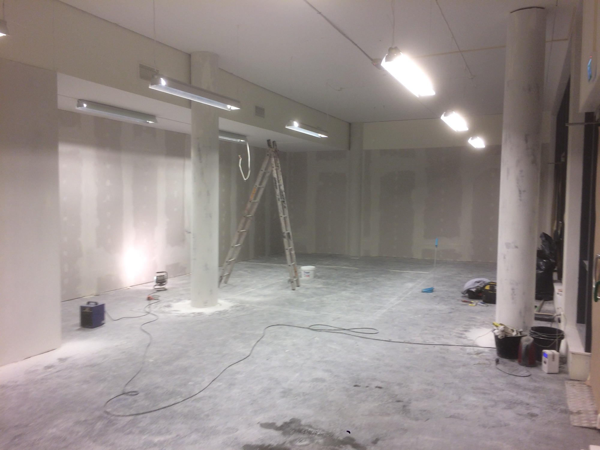Wnętrze w trakcie prac wykończeniowych: ściany pokryte gładzią, widoczna drabina, narzędzia i kable na podłodze, surowe oświetlenie.