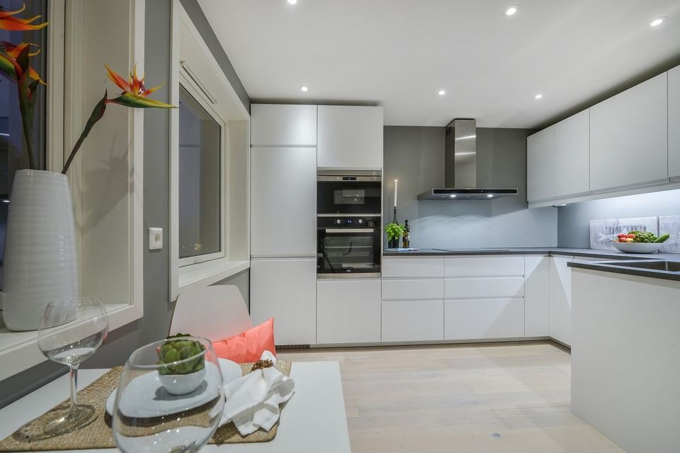 Nowoczesna, minimalistyczna kuchnia z białymi szafkami, drewnianą podłogą i kwiatami strelicji w wysokim wazonie na stole, przy oknie.