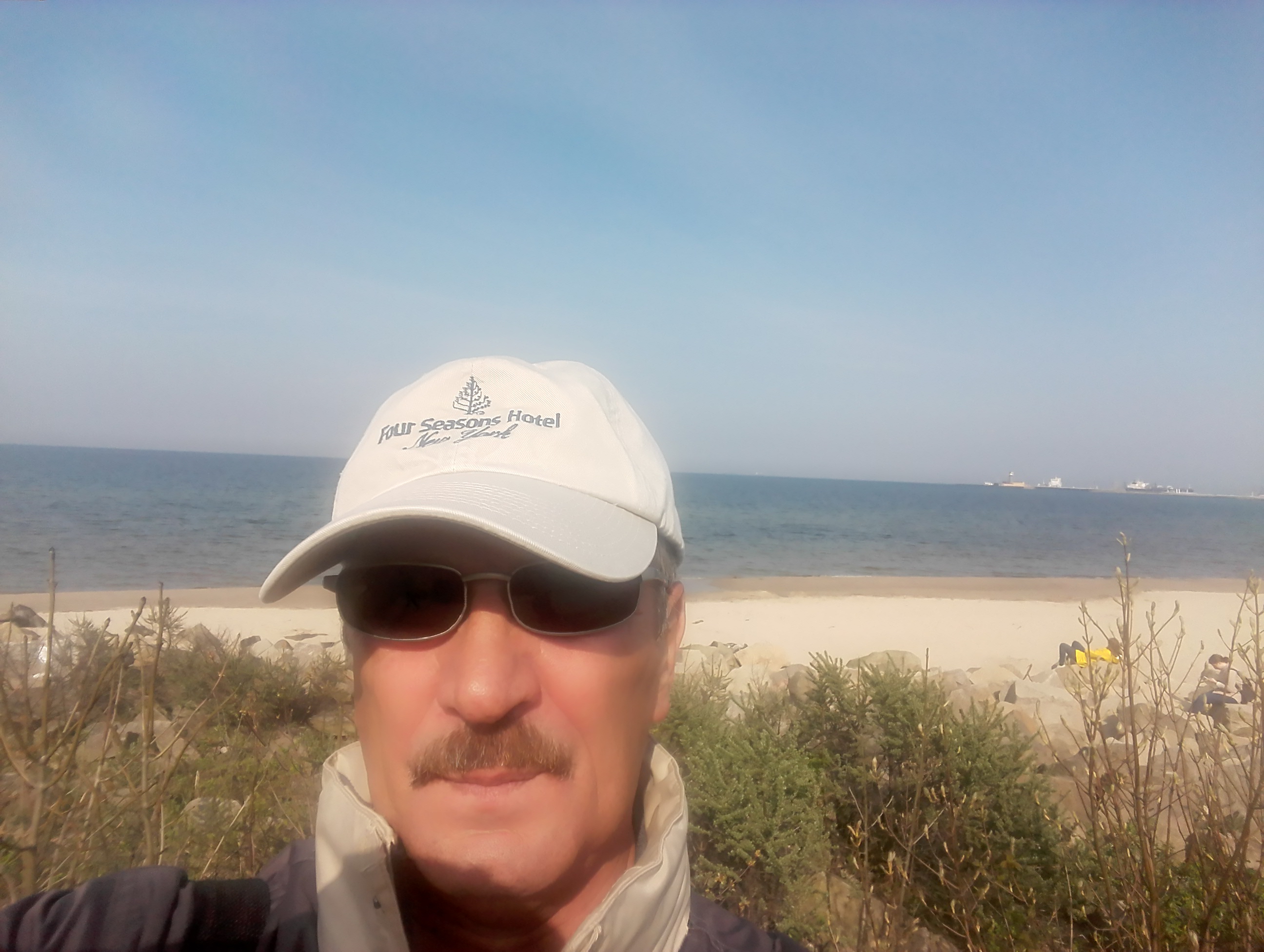 Portret mężczyzny w okularach i czapce z logo hotelu na tle morza i plaży.