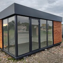 Nowoczesny pawilon biurowy z płyt warstwowych z drewnianą okładziną, szarą ramą i dużymi przeszkleniami, na żwirowym podłożu, widok z zewnątrz.