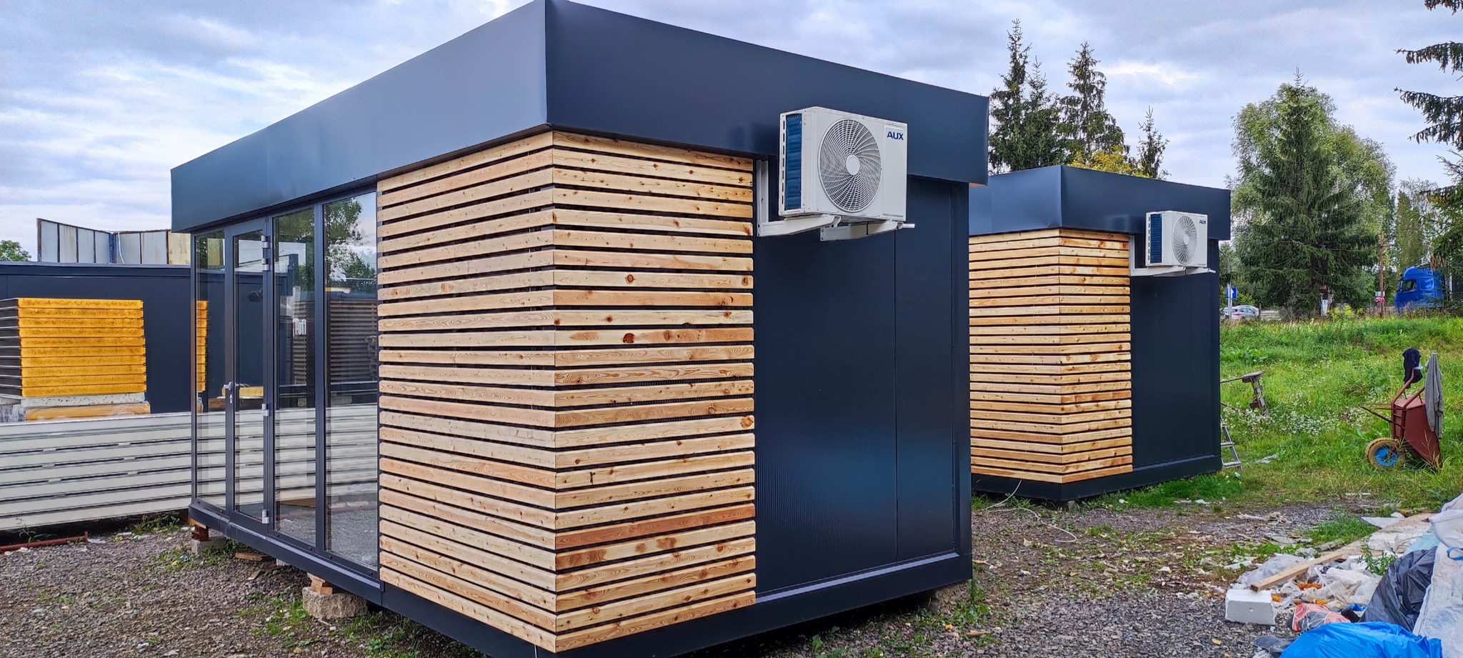 Dwa nowoczesne, modułowe pawilony z czarnymi ścianami i drewnianą okładziną, klimatyzatory zamontowane na ścianach, widoczne panele warstwowe i otoczenie budowy.