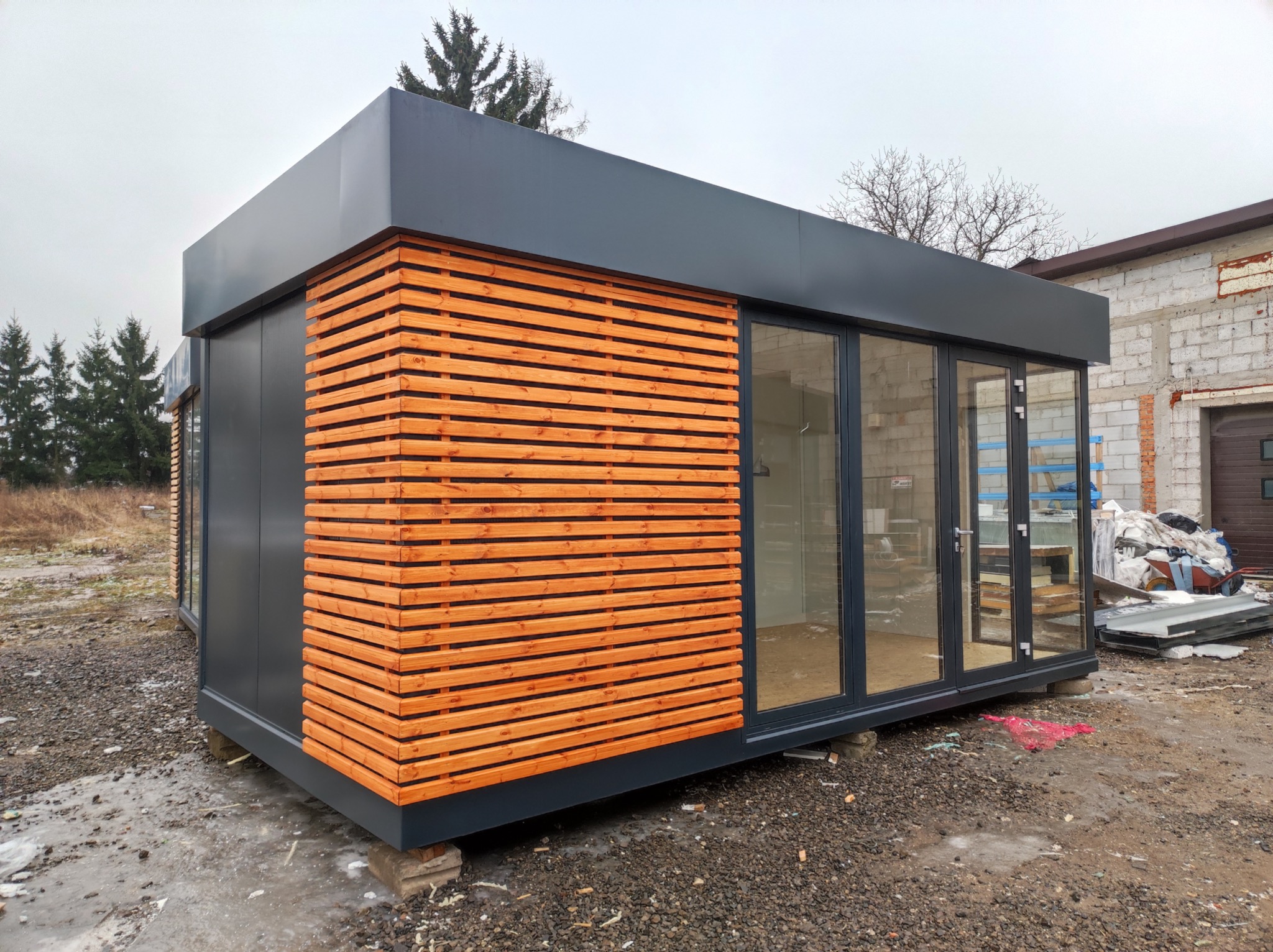 Nowoczesny pawilon modułowy z szarymi ścianami i drewnianą okładziną elewacyjną, osadzony na betonowych bloczkach na żwirowym podłożu, z widocznymi szklanymi drzwiami i oknami.