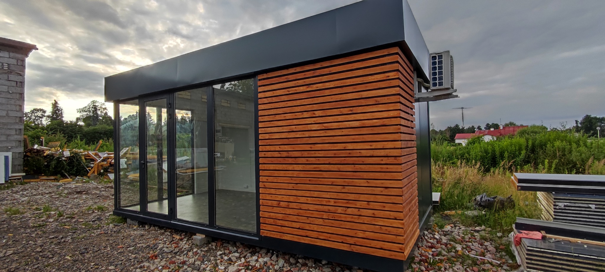 Nowoczesny pawilon handlowy z płyt warstwowych i drewnianą elewacją, z klimatyzacją na zewnątrz, na nierównym podłożu.