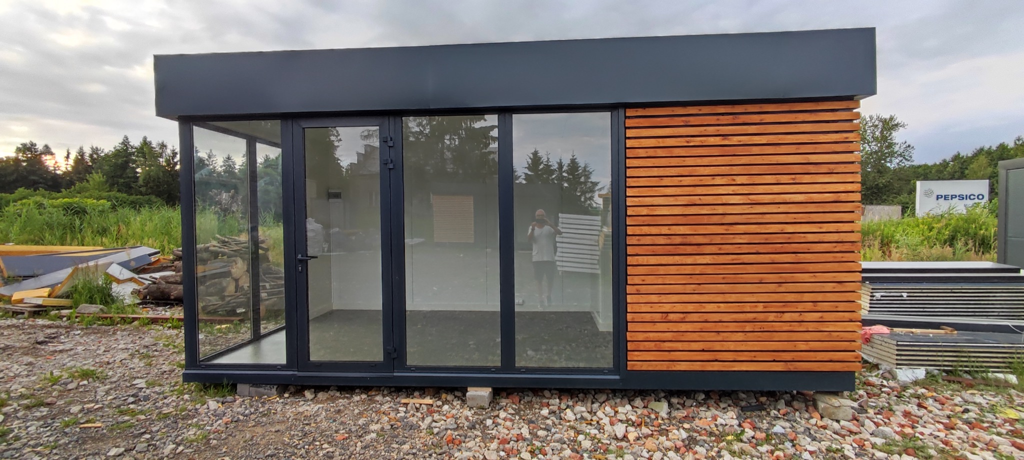 Nowoczesny pawilon z przeszklonymi ścianami i elewacją z drewnianych paneli, widoczny stos płyt warstwowych i odblask postaci w szybie.