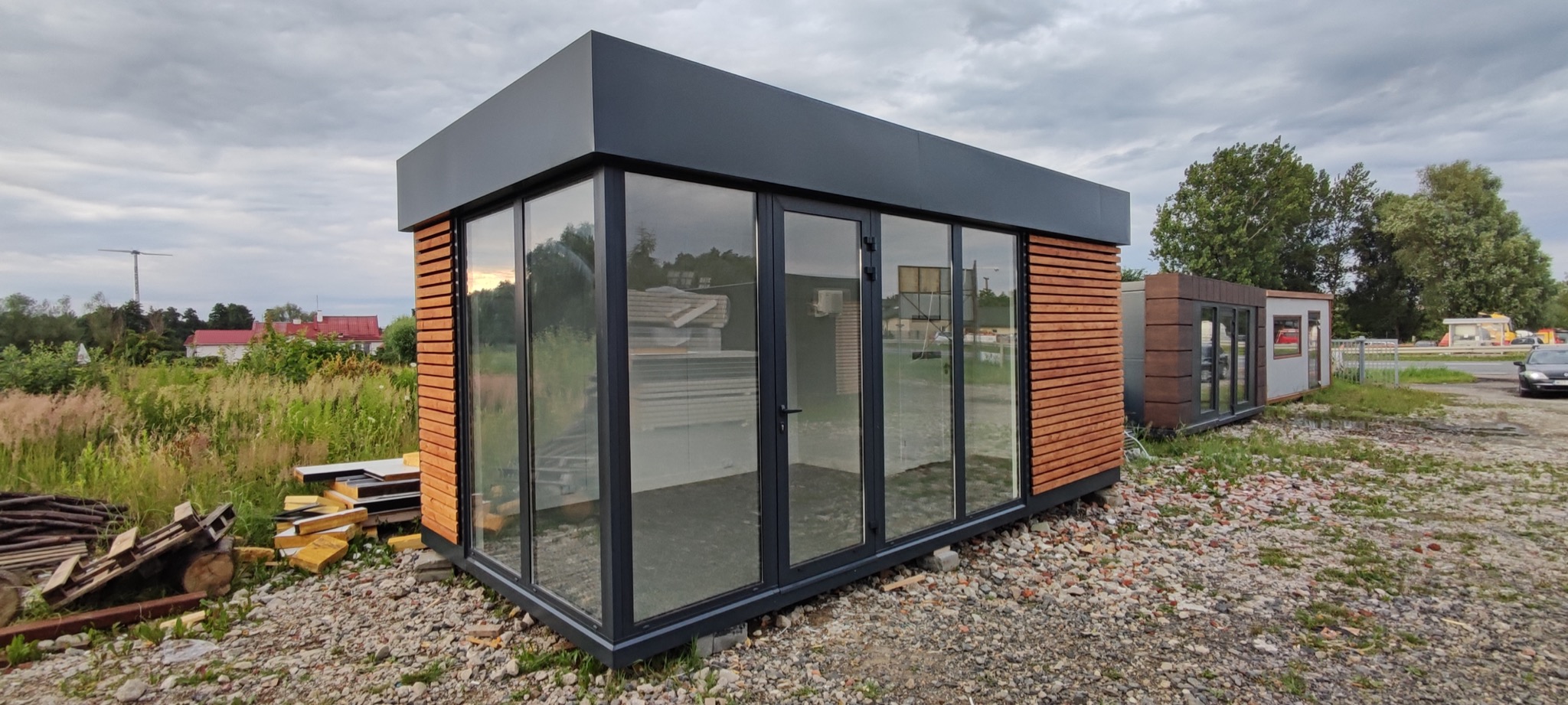 Nowoczesny pawilon biurowy z płyt warstwowych z drewnianą okładziną, szarą ramą i dużymi przeszkleniami, na żwirowym podłożu, widok z zewnątrz.