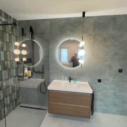 KONSILE OLEKSANDR KONONENKO - Nowoczesna łazienka z szarą, wielkoformatową płytką, kabiną prysznicową typu walk-in z czarnymi elementami, okrągłym lustrem z podświetleniem LED i wiszącą szafką z umywalką z drewnianym frontem.