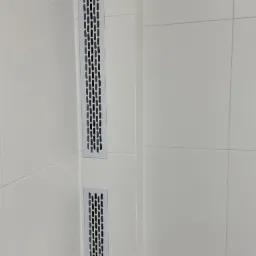 Dwa metalowe, pionowe kratki wentylacyjne wbudowane w białe płytki ceramiczne na ścianie w rogu pomieszczenia.