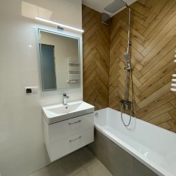 KONSILE - Nowoczesna łazienka po remoncie: biała umywalka z szafką, lustro z oświetleniem LED, wanna z drewnopodobną obudową i deszczownicą. Minimalistyczny design.