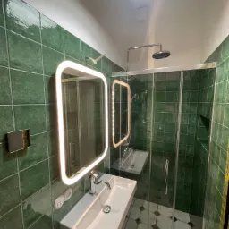 Nowoczesna łazienka z zielonymi, kwadratowymi płytkami, kabiną prysznicową ze szklanymi drzwiami i podświetlanym lustrem nad białą umywalką. Widoczne okrągłe okno w suficie.