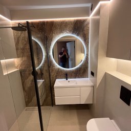 KONSILE - Nowoczesna łazienka z beżowymi płytkami i marmurową ścianą, okrągłym lustrem LED, podwieszaną szafką z umywalką, kabiną walk-in z czarną ramą i deszczownicą.