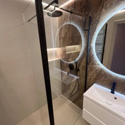 KONSILE - Nowoczesna łazienka z kabiną walk-in o czarnej ramie, deszczownicą i liniowym odpływem. Okrągłe lustro LED nad białą szafką z umywalką, ściany z marmurowymi i beżowymi płytkami, oświetlenie liniowe.