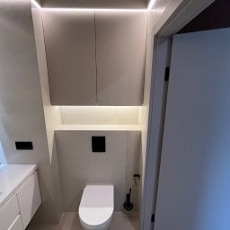KONSILE - Minimalistyczna łazienka z podwieszaną toaletą, beżowymi szafkami w zabudowie i subtelnym oświetleniem LED. Widoczna biała szafka umywalkowa.