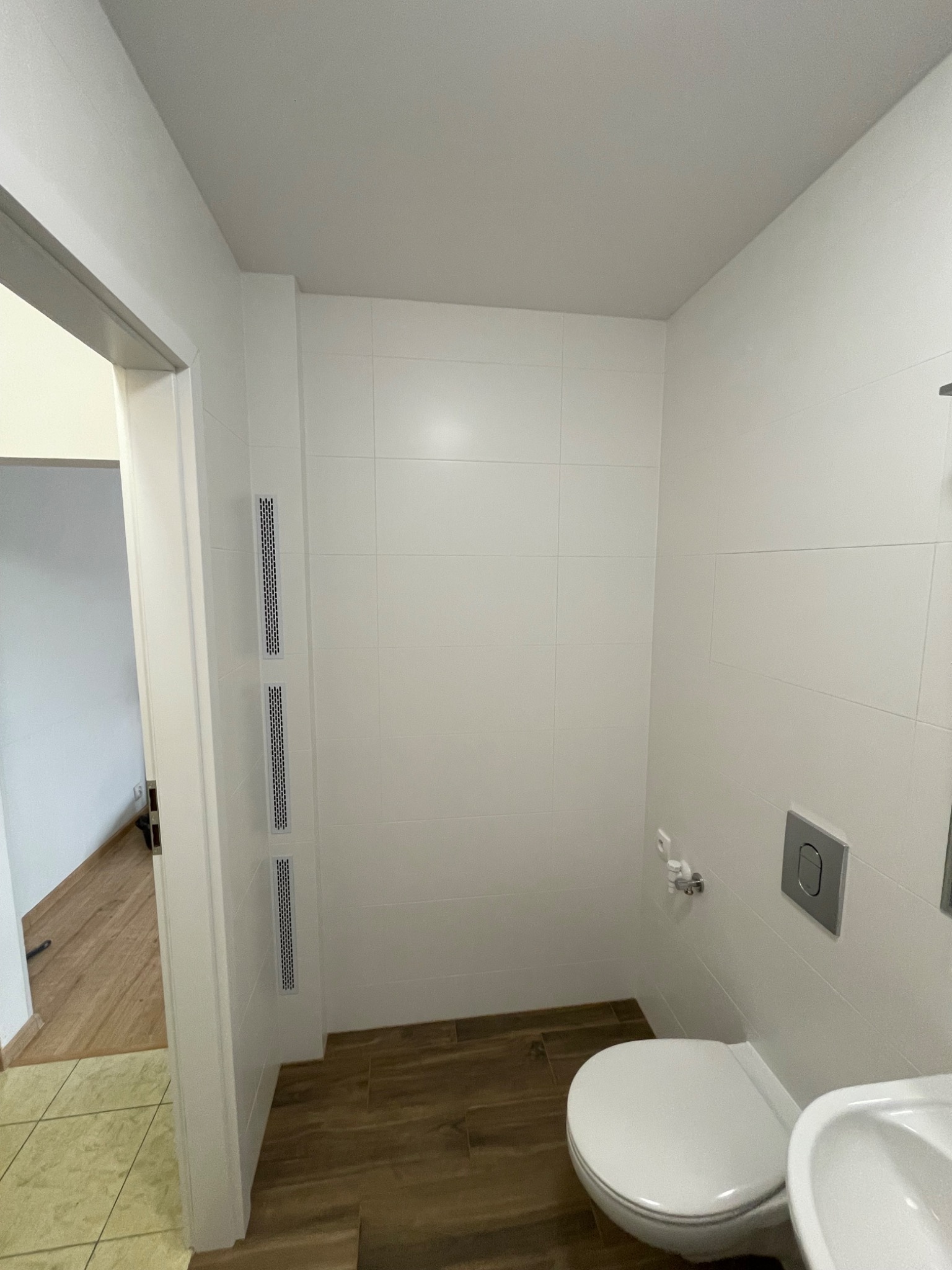 Nowoczesna, minimalistyczna toaleta z białymi płytkami, drewnopodobną podłogą, podwieszaną miską WC z chromowanym przyciskiem, oraz trzema pionowymi kratkami wentylacyjnymi w ścianie obok drzwi.