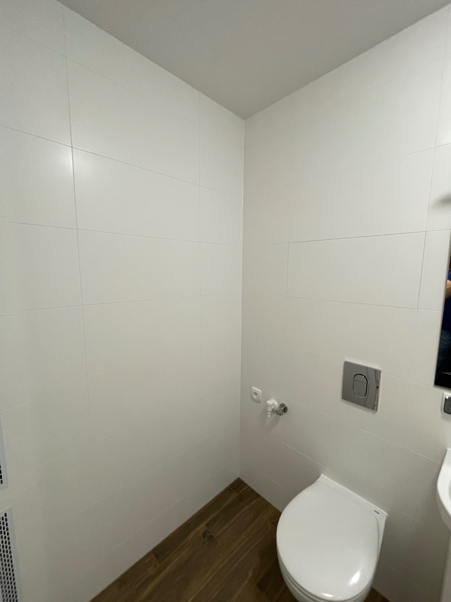 Wnętrze małej, nowoczesnej toalety z białymi płytkami ściennymi, drewnopodobną podłogą, podwieszaną muszlą klozetową i minimalistycznym przyciskiem spłukującym. Widoczna instalacja wodna i fragment...