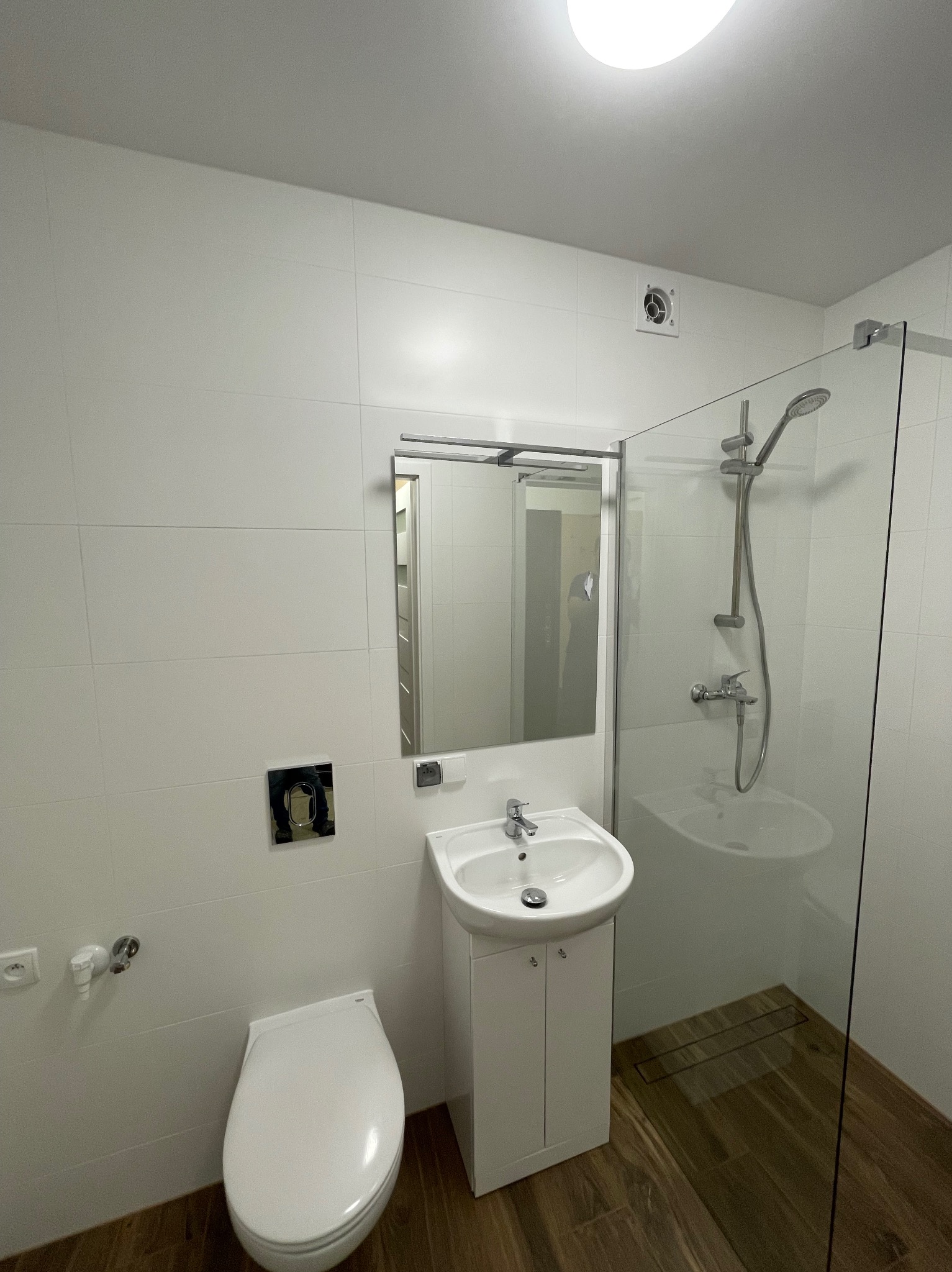 Nowoczesna, minimalistyczna łazienka z białymi płytkami, drewnopodobną podłogą, kabiną prysznicową ze szklaną ścianką, umywalką na szafce i podwieszaną toaletą z chromowanym przyciskiem.