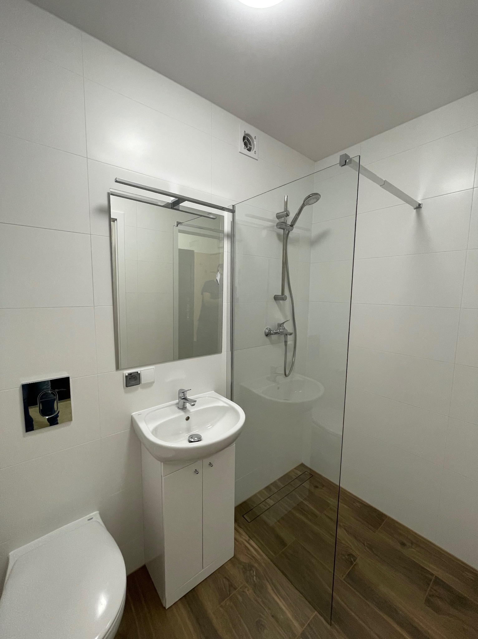 Nowoczesna łazienka z białymi płytkami, kabiną prysznicową ze szklaną ścianką, umywalką na szafce i drewnopodobną podłogą z odpływem liniowym.