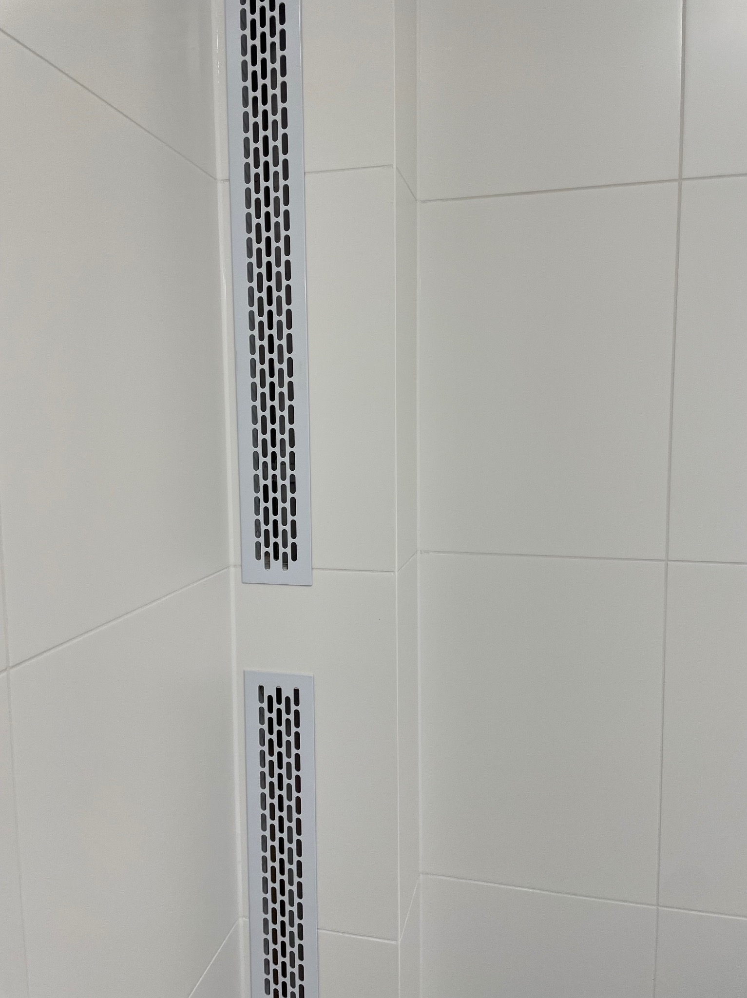 Dwa metalowe, pionowe kratki wentylacyjne wbudowane w białe płytki ceramiczne na ścianie w rogu pomieszczenia.
