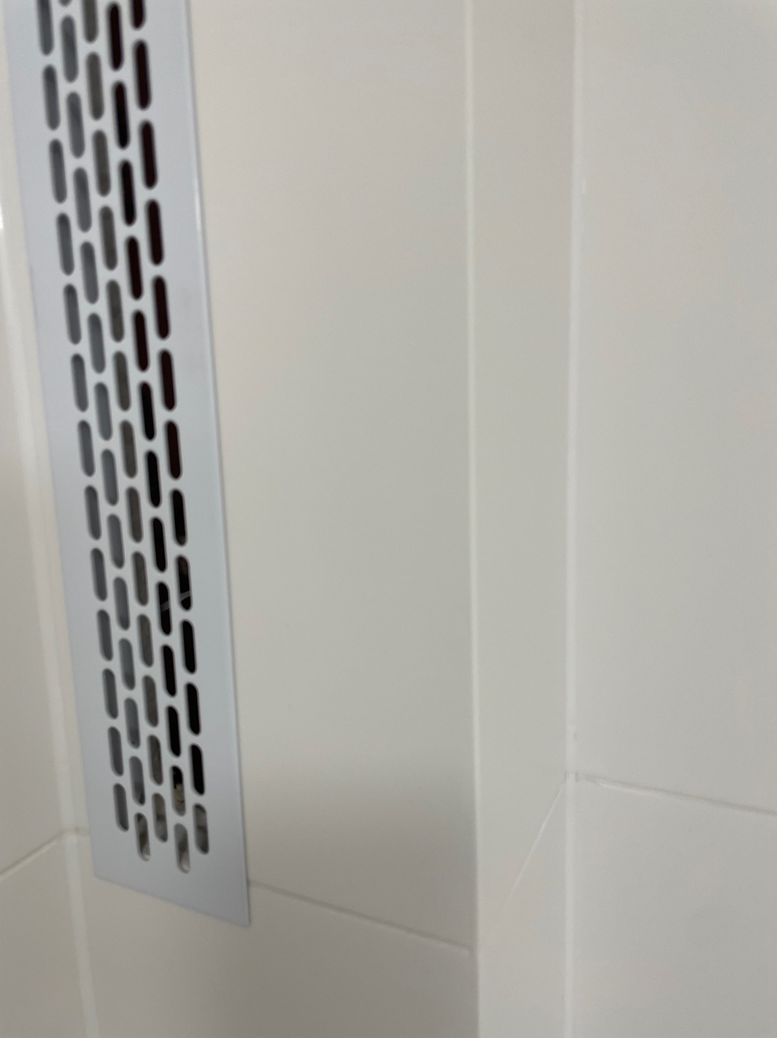 Białe płytki ceramiczne w łazience z metalową kratką wentylacyjną wbudowaną w ścianę, widoczne fugi i narożnik. Minimalistyczny design.