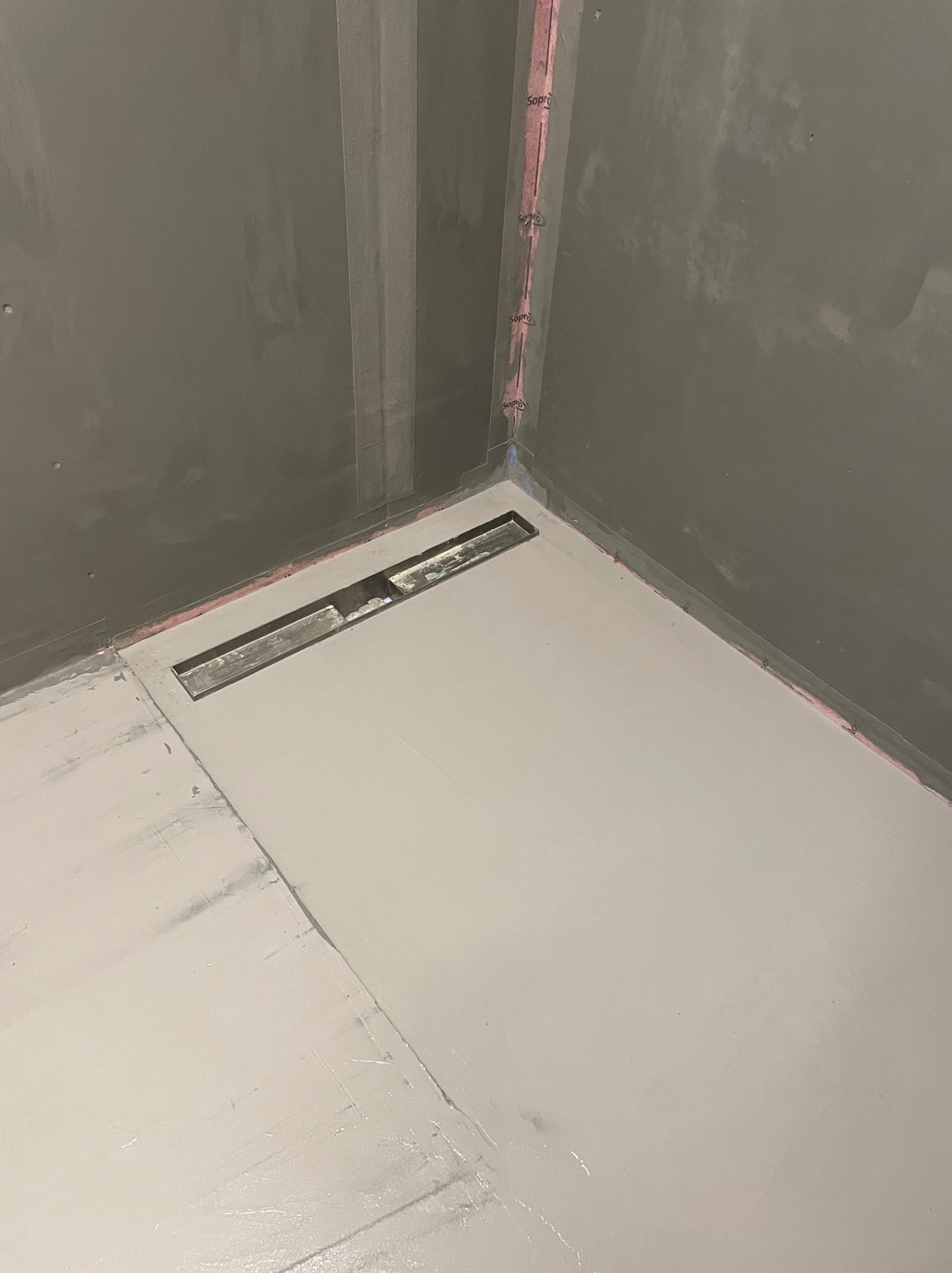 Przygotowana do dalszych prac powierzchnia podłogi w łazience z zamontowanym odpływem liniowym. Ściany przygotowane pod płytki, zabezpieczone różową taśmą uszczelniającą.