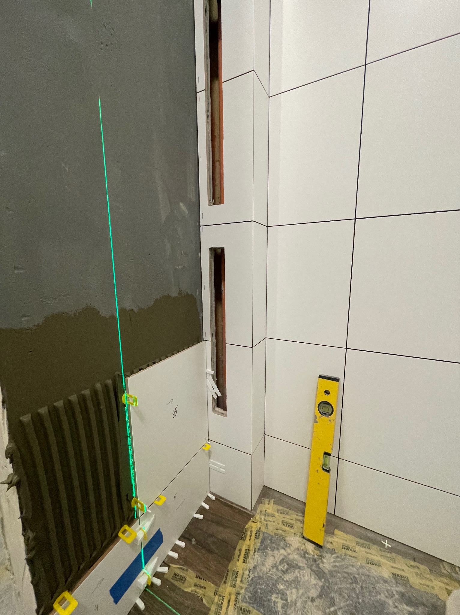 Układanie białych płytek kwadratowych w łazience, z użyciem lasera krzyżowego i poziomicy; widoczne szpachelki dystansowe i fragment surowej ściany z nałożonym klejem.