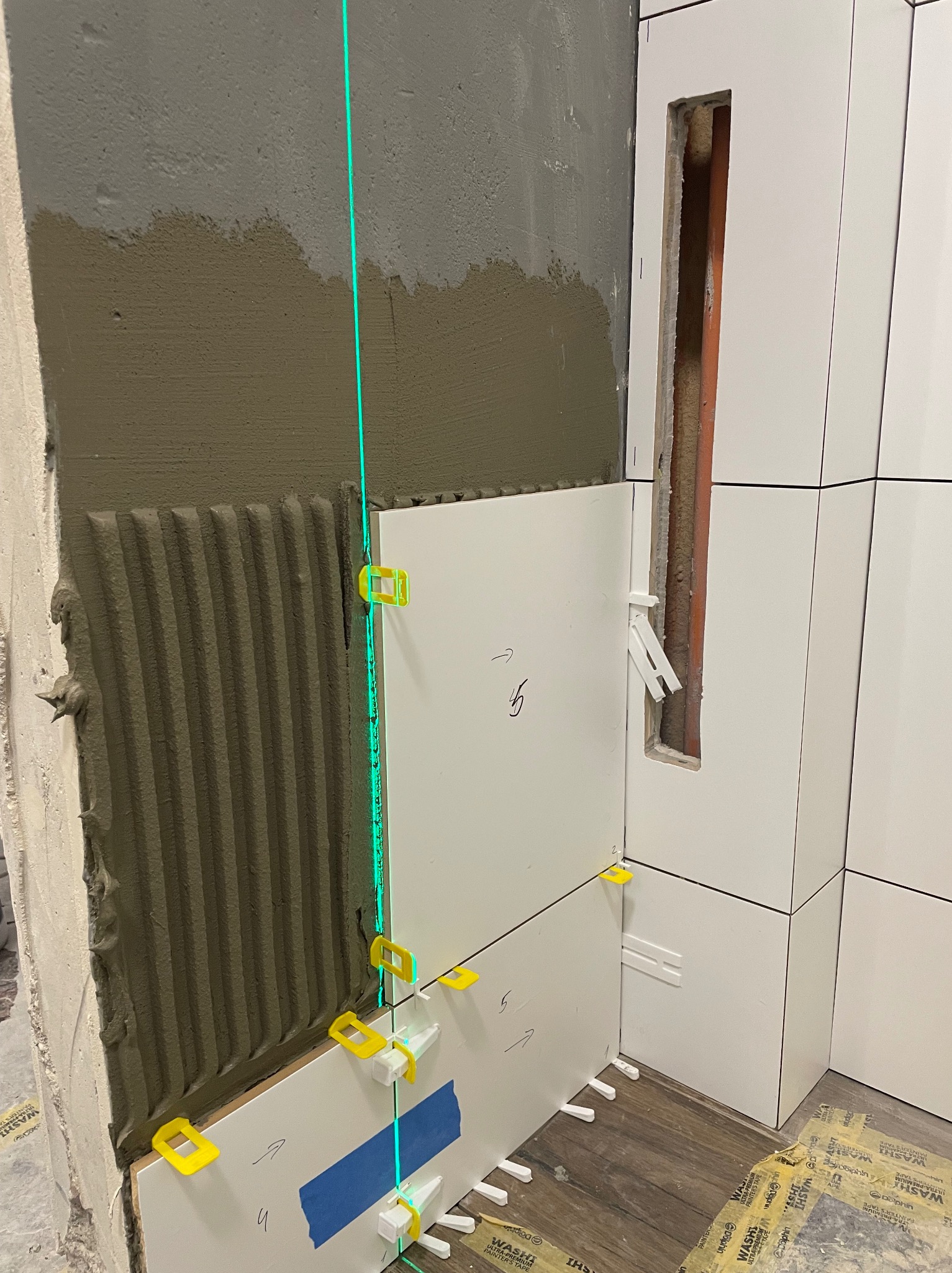 Układanie białych płytek ściennych z użyciem poziomicy laserowej i klipsów poziomujących, widoczne ślady zaprawy na ścianie i taśma malarska.