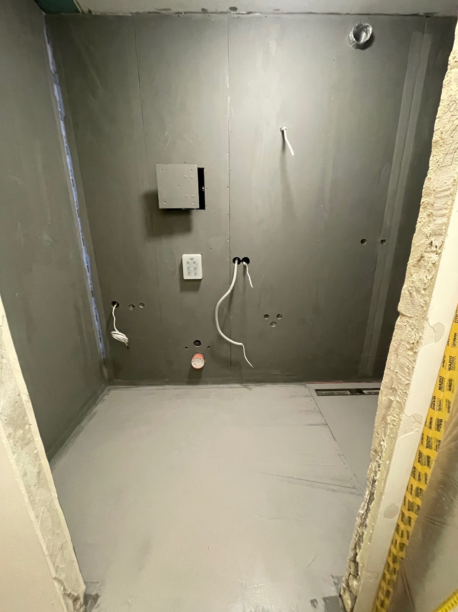 Surowa łazienka w trakcie przygotowania do dalszych prac, ściany pokryte szarą warstwą izolacyjną, widoczne wystające kable i otwory na instalacje, podłoga zabezpieczona folią, fragment futryny...