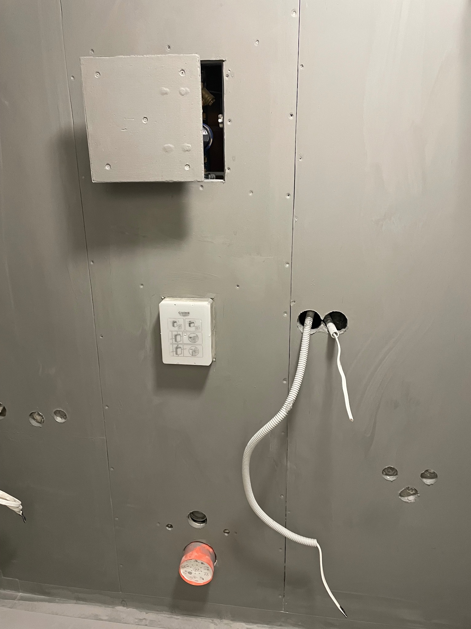 Szara ściana z płyt kartonowo-gipsowych w łazience, z widocznymi otworami na instalacje, puszką rewizyjną z otwartymi drzwiczkami, białą puszką instalacyjną Grohe i wystającymi przewodami.