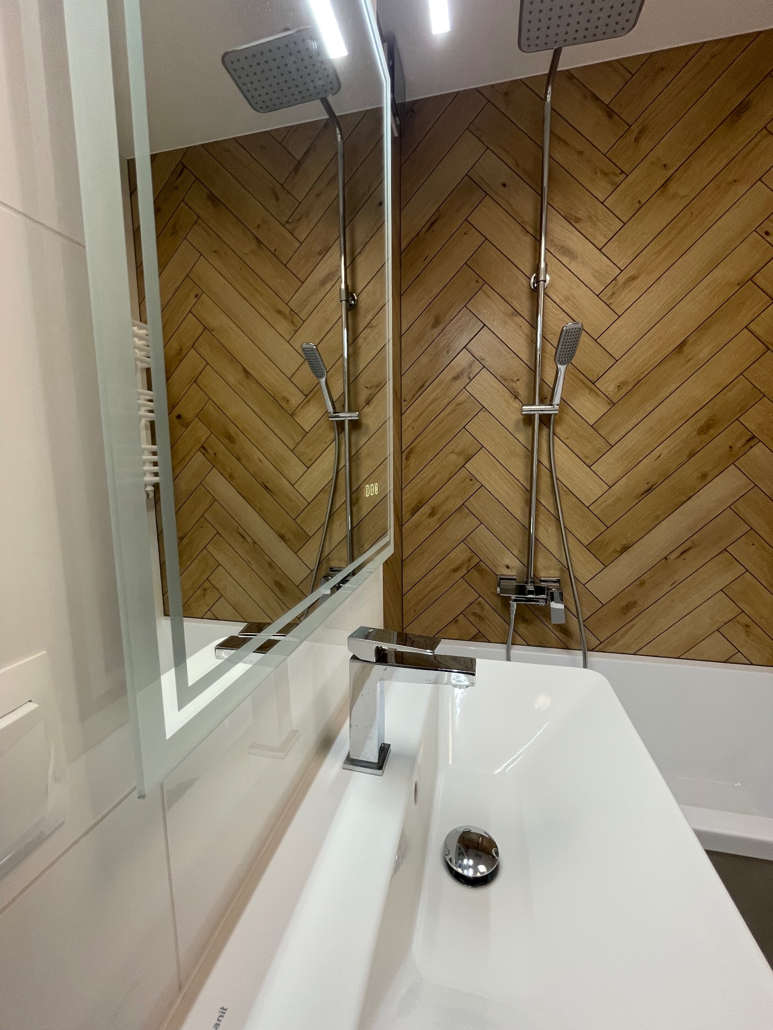 Nowoczesna łazienka z drewnopodobną ścianą w jodełkę, chromowaną armaturą i podświetlanym lustrem. Umywalka z baterią w minimalistycznym stylu.