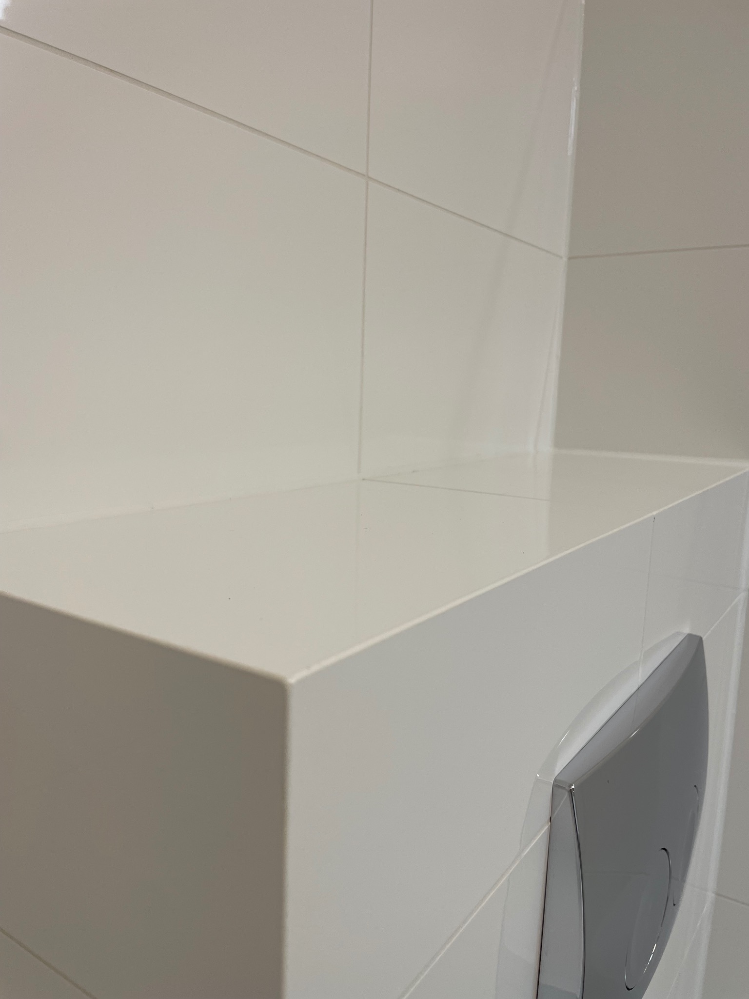 Biała, błyszcząca zabudowa WC z płytkami ceramicznymi, chromowany przycisk spłukujący. Minimalistyczny design, czyste linie, nowoczesna łazienka po remoncie.