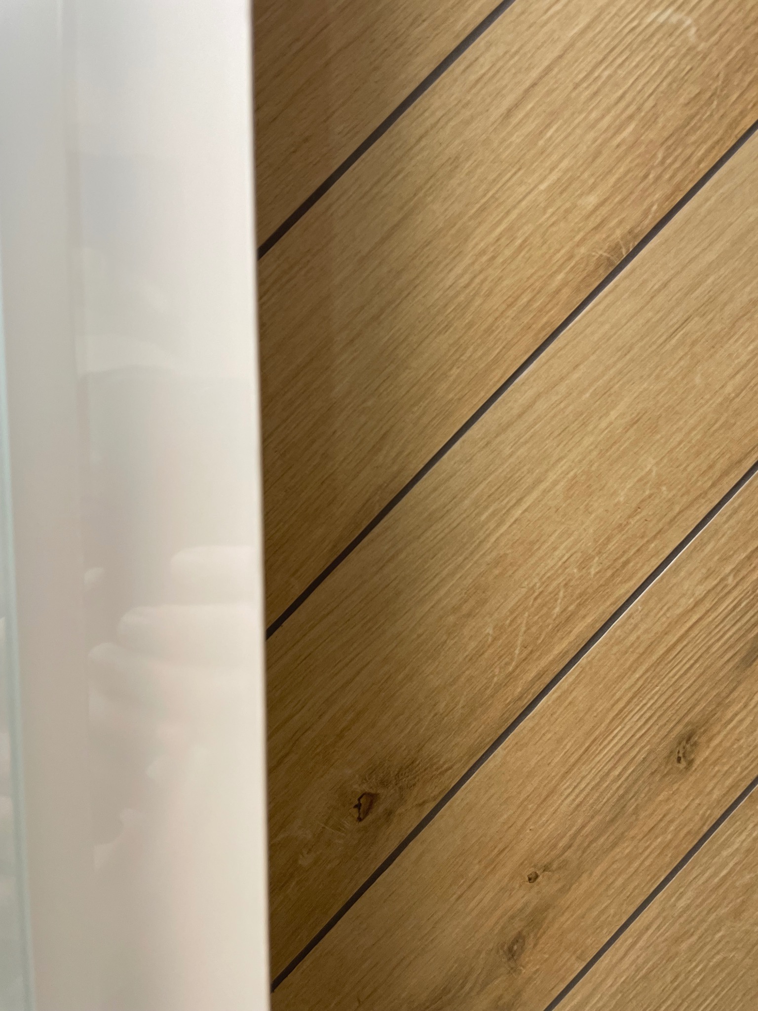 Ukończona ściana z płytek imitujących drewno, ułożonych pod kątem, z fragmentem białej armatury łazienkowej po lewej stronie. Detal wykończenia łazienki.