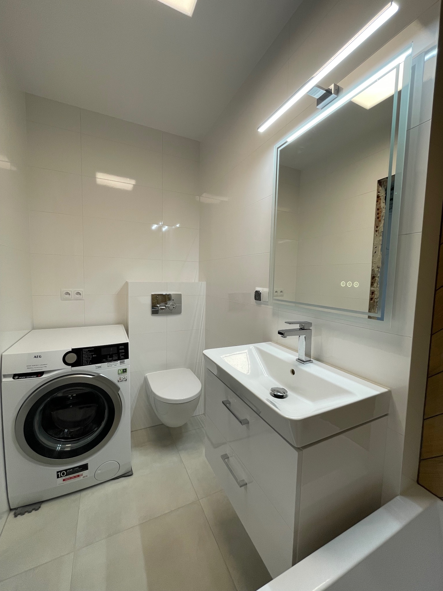 Nowoczesna, jasna łazienka z białą armaturą: umywalka z szafką, toaleta podwieszana, pralka AEG. Minimalistyczne płytki ścienne i podłogowe, lustro z podświetleniem LED.