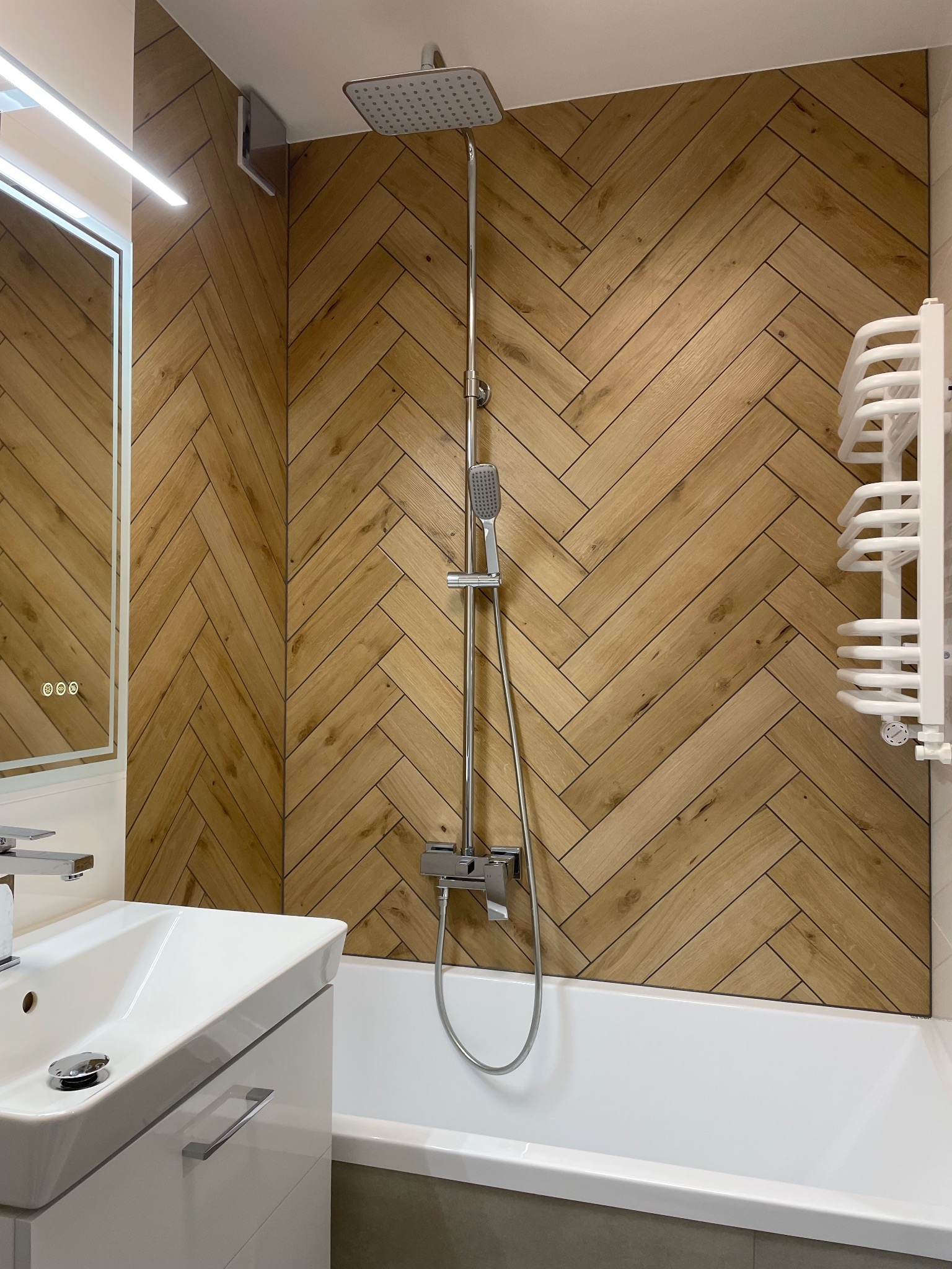Nowoczesna łazienka z drewnopodobnymi płytkami ułożonymi w jodełkę na ścianie, białą wanną i umywalką z szafką. Chromowana armatura i podświetlane lustro.