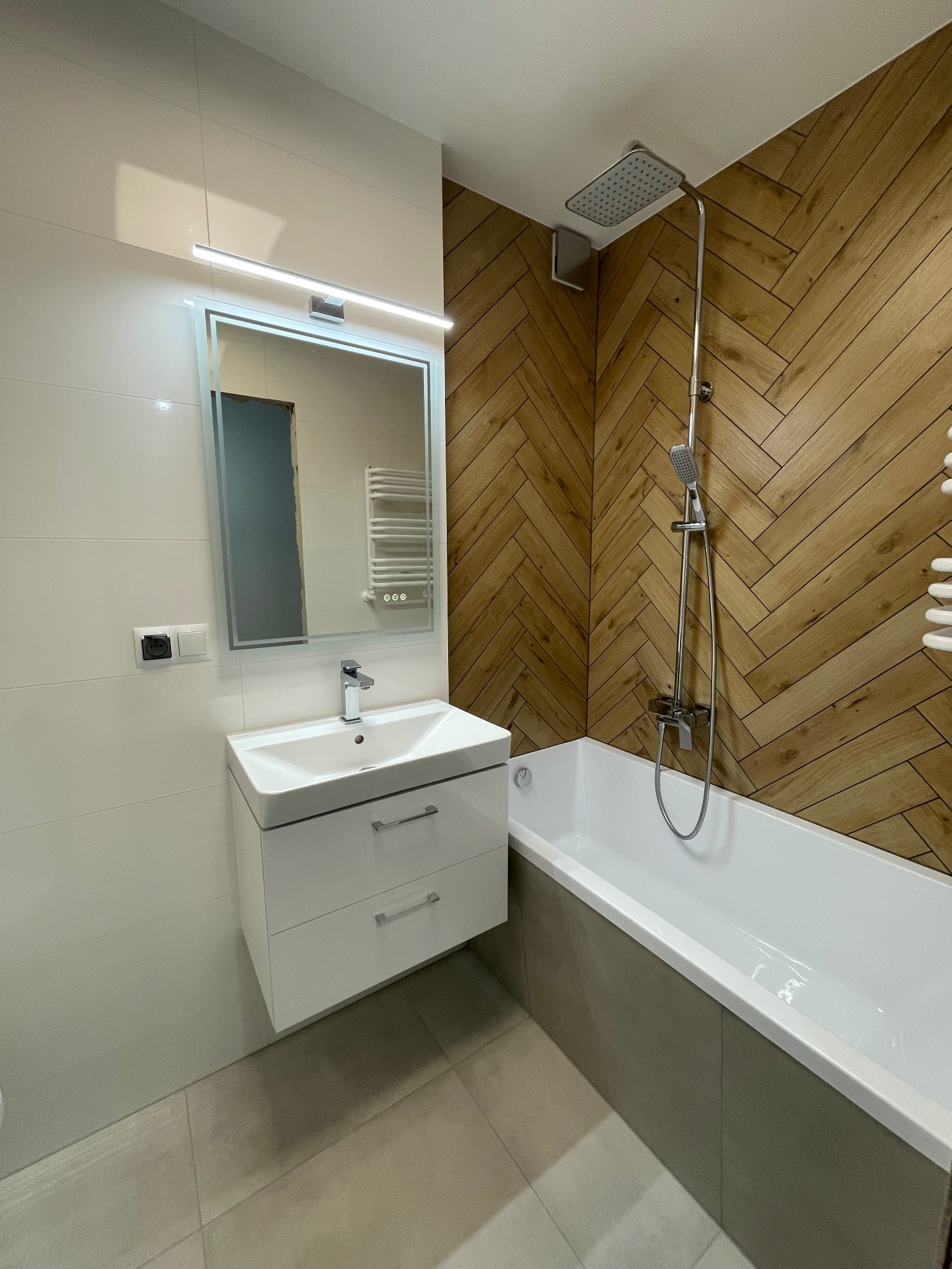 Nowoczesna łazienka po remoncie: biała umywalka z szafką, lustro z oświetleniem LED, wanna z drewnopodobną obudową i deszczownicą. Minimalistyczny design.