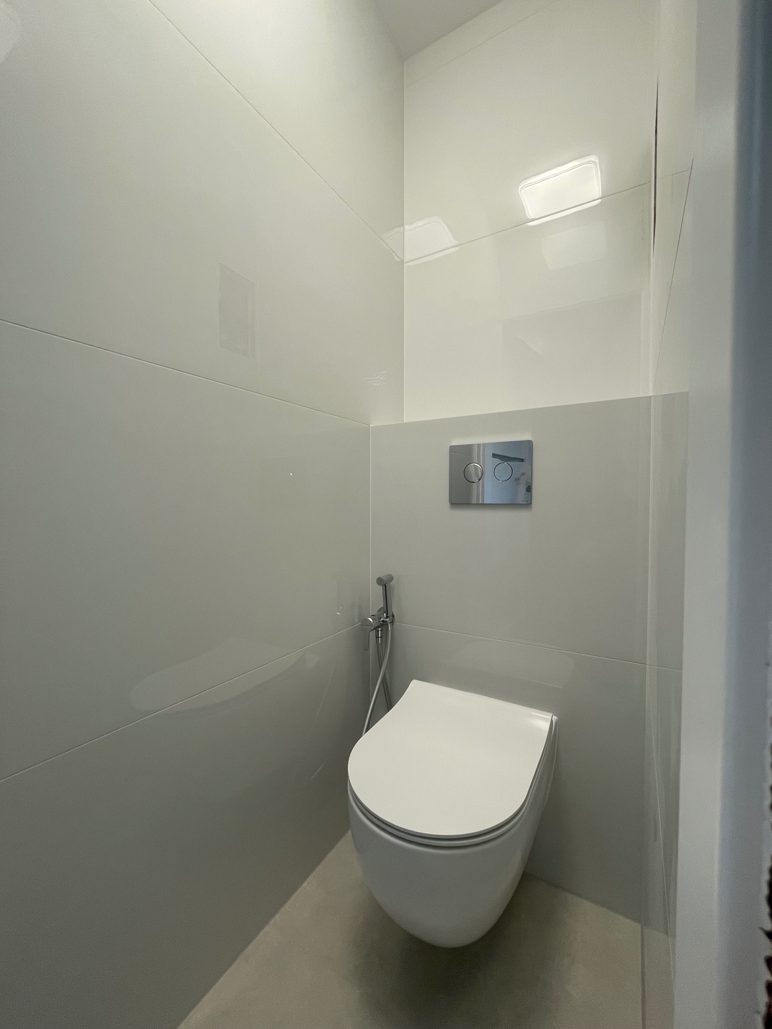 Minimalistyczna toaleta podwieszana z bidetką i chromowanym przyciskiem spłukującym, wykończona jasnymi, błyszczącymi płytkami w nowoczesnej łazience.