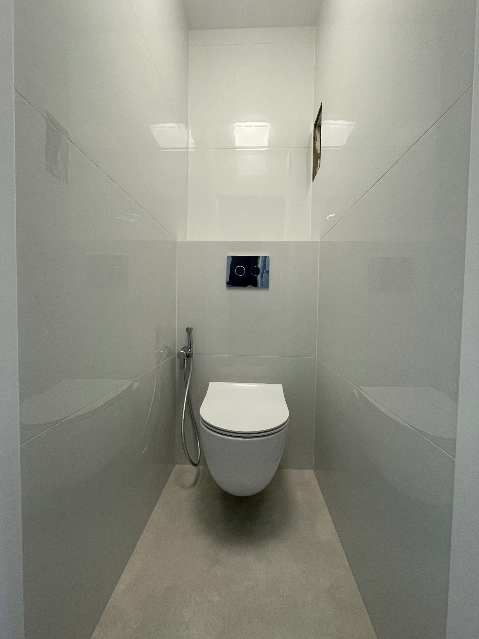 Minimalistyczna toaleta w wąskiej, wykafelkowanej na biało wnęce. Podwieszana muszla klozetowa, szara podłoga, chromowana bateria bidetowa. Nowoczesny design.